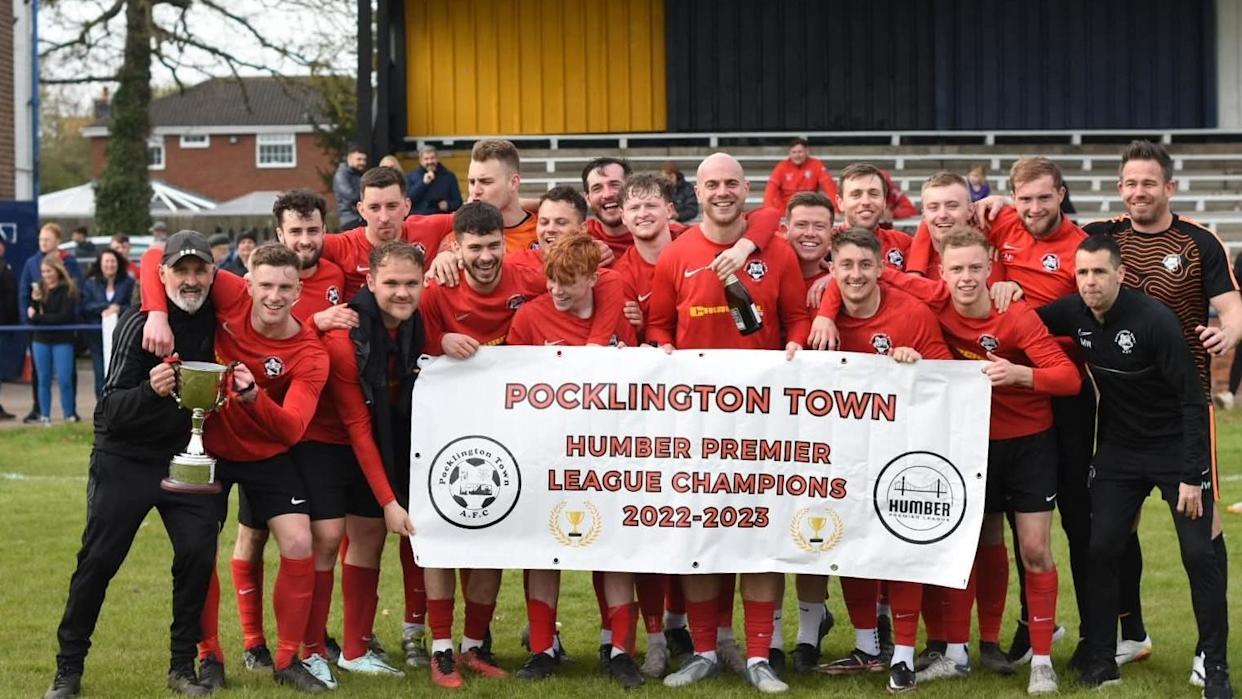 The team stand in red socks and jerseys and two players are holding up the silver cup.  Others hold a sign that reads Pocklington Town, Humber Premier League Champions 2022-2023.