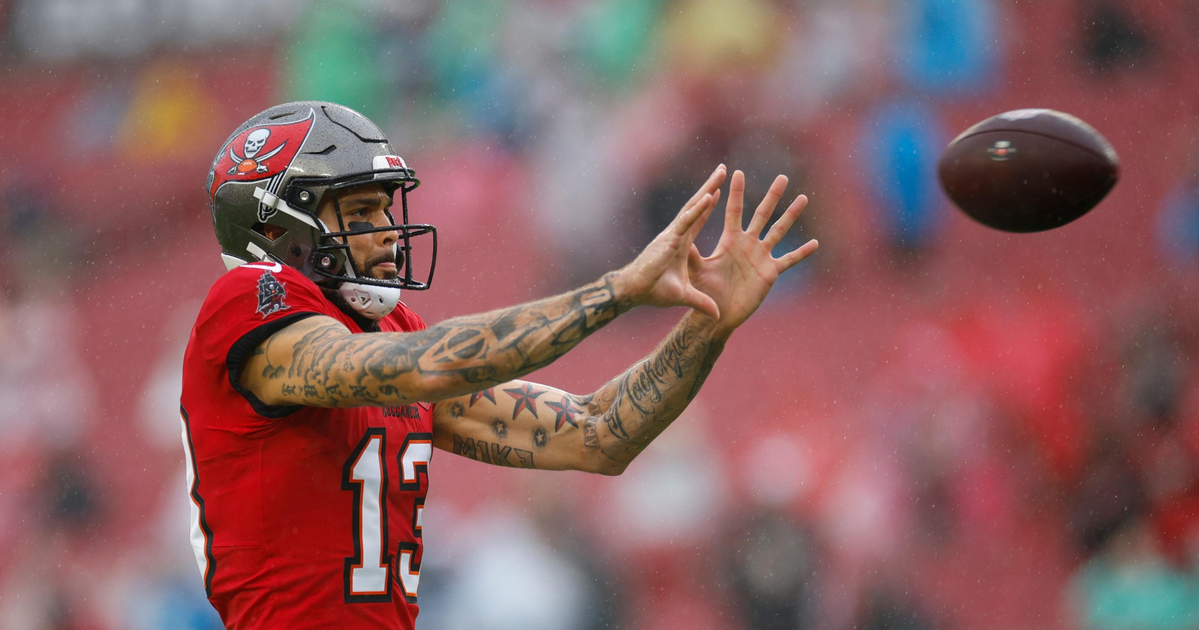 A football player wearing a red Tampa Bay Buccaneers jersey and helmet reaches out with both hands to catch a football in mid-air.