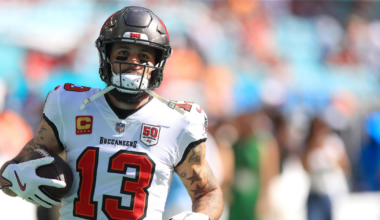 A football player in a white Tampa Bay Buccaneers jersey with number 13 holds the ball, running on a field with a blurred crowd in the background.