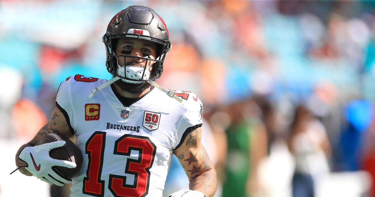 A football player in a white Tampa Bay Buccaneers jersey with number 13 holds the ball, running on a field with a blurred crowd in the background.