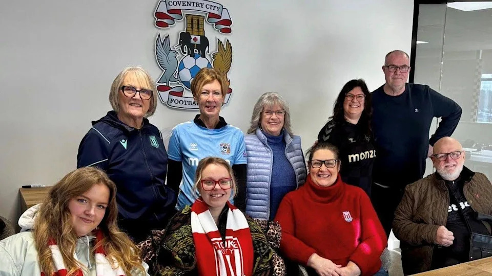 A group of six women and two men all of different ages are arranged in a row of two, the front row is sitting down and the back row are standing behind them. In the background on a white wall is a Coventry city logo