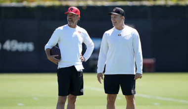 Two men stand on a grassy field, wearing similar sports attire: white long-sleeve tops, black shorts, and caps. One holds a football, and they appear to be smiling.