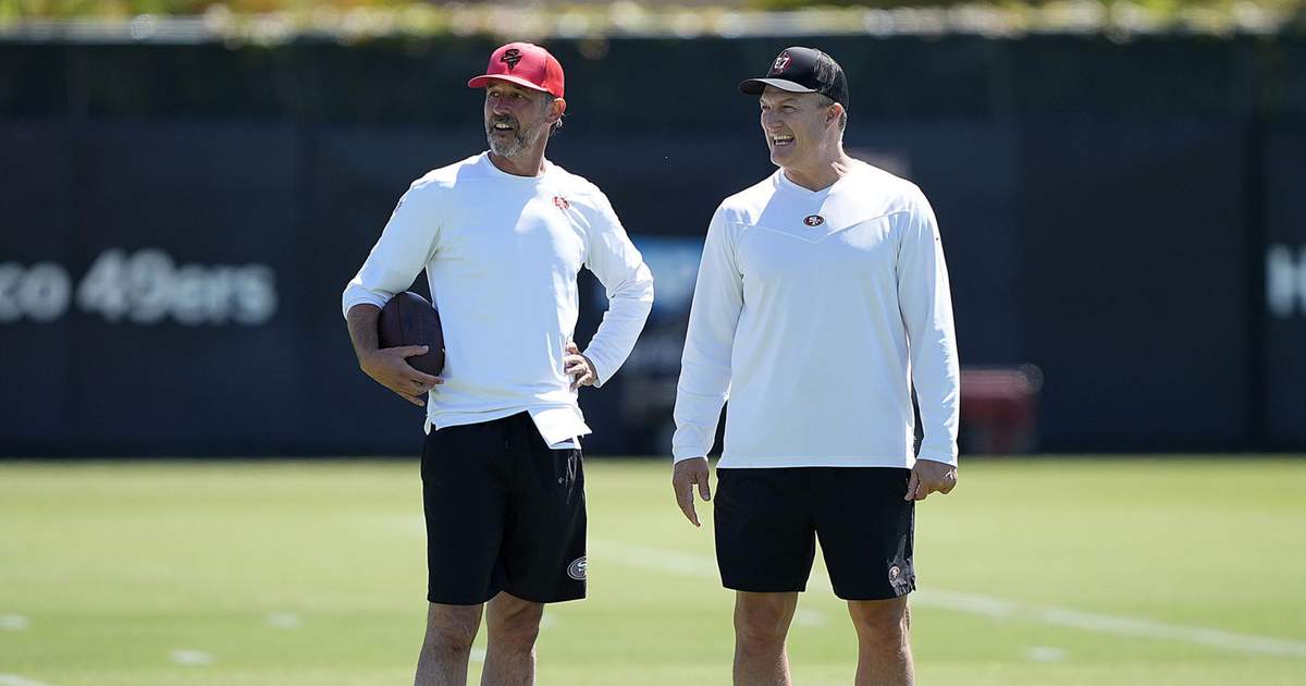 Two men stand on a grassy field, wearing similar sports attire: white long-sleeve tops, black shorts, and caps. One holds a football, and they appear to be smiling.