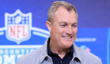 A middle-aged man in a gray jacket and black shirt smiles while speaking at an NFL Scouting Combine event.