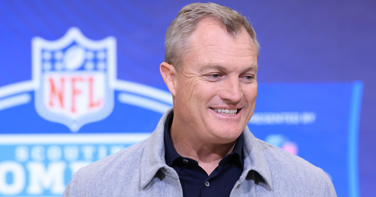 A middle-aged man in a gray jacket and black shirt smiles while speaking at an NFL Scouting Combine event.