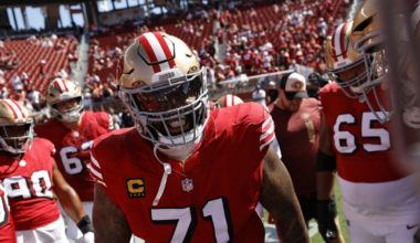 A football player wearing a red jersey with number 71 and gold helmet stands among teammates in a stadium filled with fans.