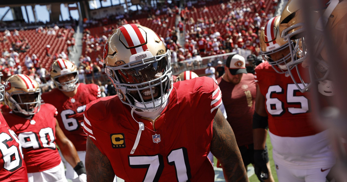 A football player wearing a red jersey with number 71 and gold helmet stands among teammates in a stadium filled with fans.
