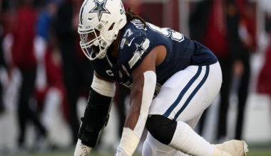 A Dallas Cowboys football player in full gear crouches on the field, ready to start a play, with blurred spectators in the background.