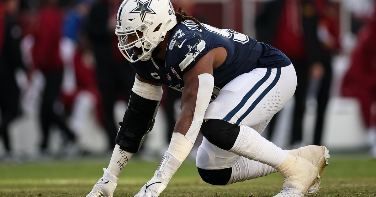 A Dallas Cowboys football player in full gear crouches on the field, ready to start a play, with blurred spectators in the background.