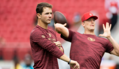 Two men wearing matching maroon long-sleeve shirts practice football throws on a field with red stadium seats in the background.