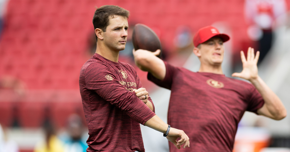 Two men wearing matching maroon long-sleeve shirts practice football throws on a field with red stadium seats in the background.