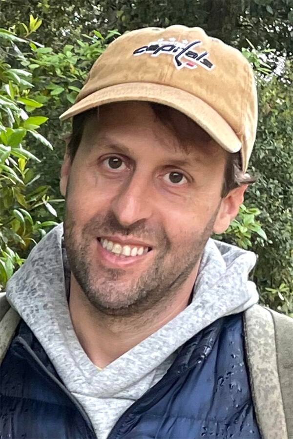 A headshot of David Covucci, a light-skinned man wearing a Washington Capitals baseball cap and a gray hoodie.