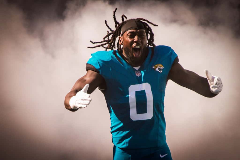 Devin Lloyd emerges through smoke during player introductions, wearing a teal Jacksonville Jaguars jersey with number 0, shouting with intensity and arms extended as he runs onto the field.