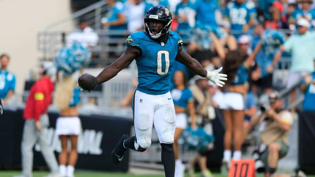 Devin Lloyd celebrates during a Jacksonville Jaguars game, holding the football in his right hand while spreading his arms wide in a teal number 0 jersey as the crowd looks on in the background.