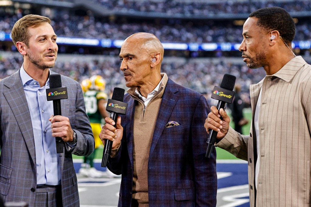 Three male sports commentators hold NBC Sports microphones at a football game.