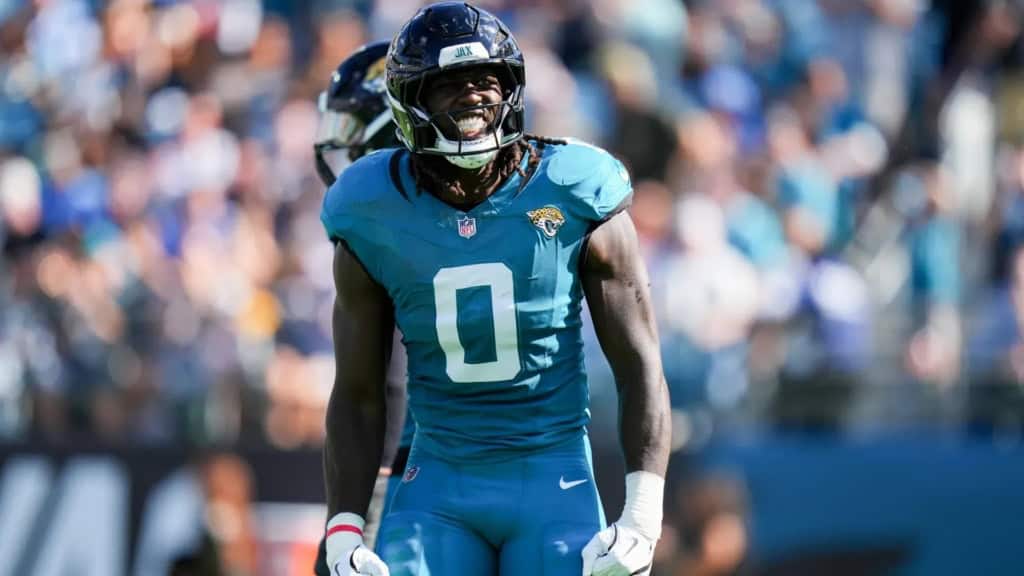 Devin Lloyd stands on the field in a teal Jacksonville Jaguars number 0 uniform, flexing and smiling intensely after a play with the stadium crowd blurred behind him.
