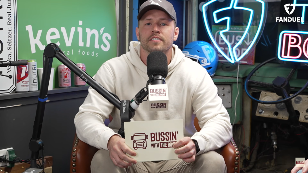 A man wearing a hat and hoodie speaks into a microphone that has "Bussin' with the Boys" on it, while holding a card with the same logo.