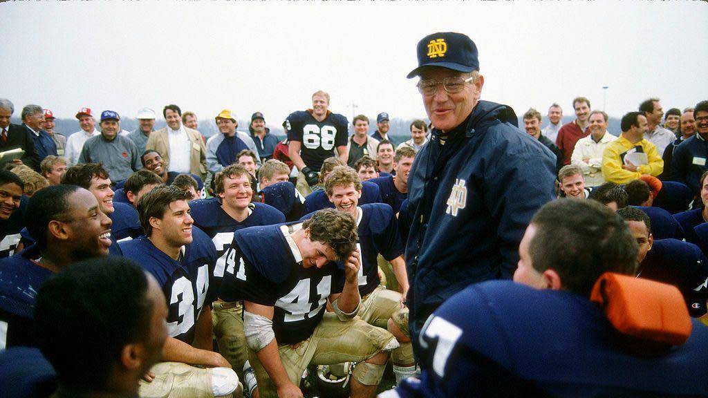 Lou Holtz standing at the centre of a group of kneeling players, and speaking. He's in a baseball cap and windbreaker with the Notre Dame logo 