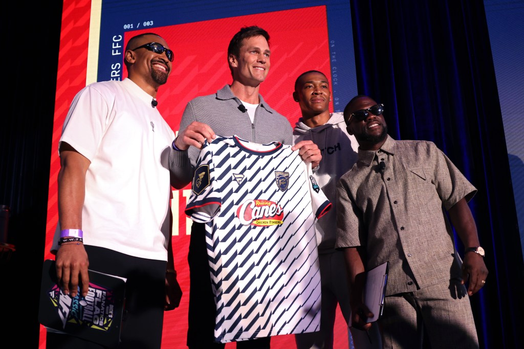Four men, including Tom Brady and Kevin Hart, on a stage with a blue and white striped jersey for the Fanatics Flag Football Classic.