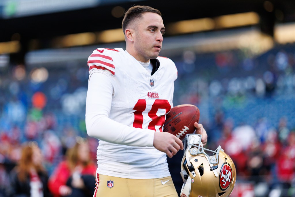 Eddy Pineiro #18 of the San Francisco 49ers stands on the field holding a football and a helmet.