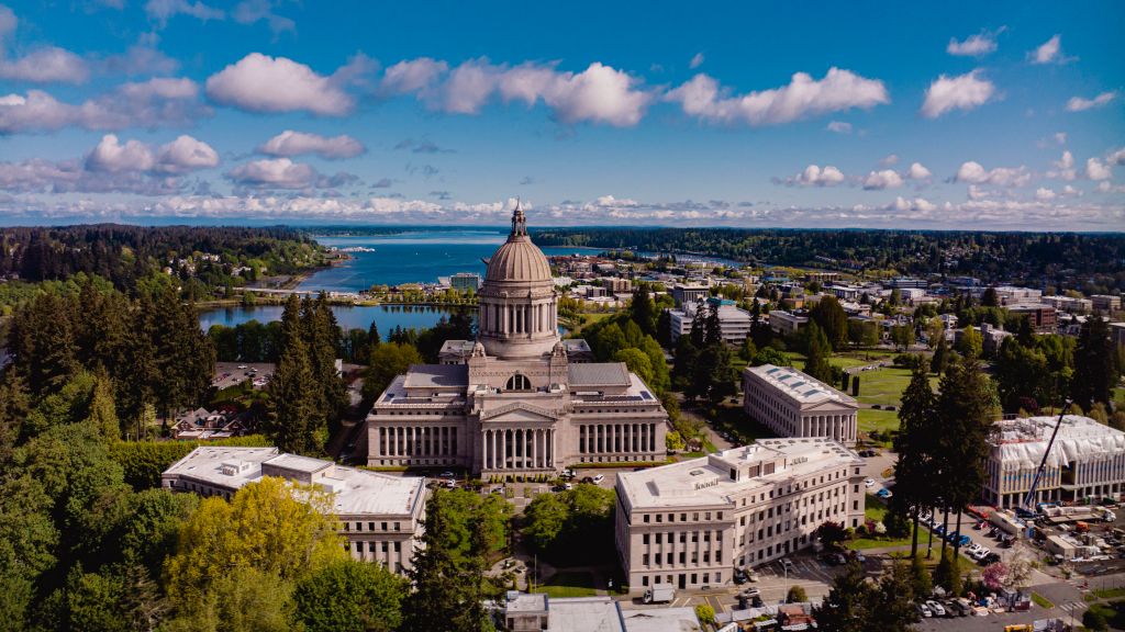 Aerial view of the Washington State Capitol building and surrounding city, with a body of water and distant mountains under a blue sky with scattered clouds.