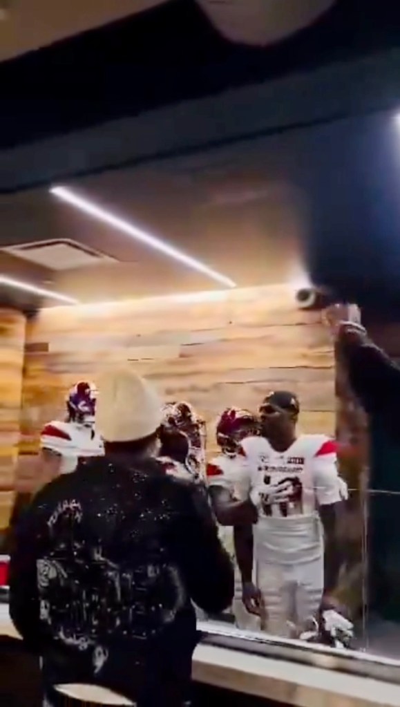 Two men in athletic uniforms behind a window in a restaurant, a man with a beanie and black sweatshirt in front of the window, and a hand holding up a camera.