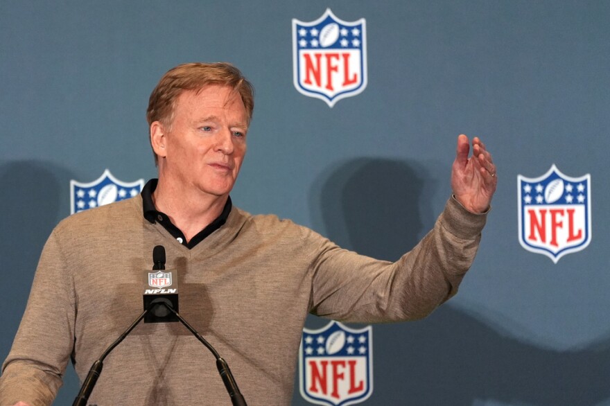 Man in light brown sweater gestures with hand while speaking into microphone. Backdrop is blue with multiple NFL shields 