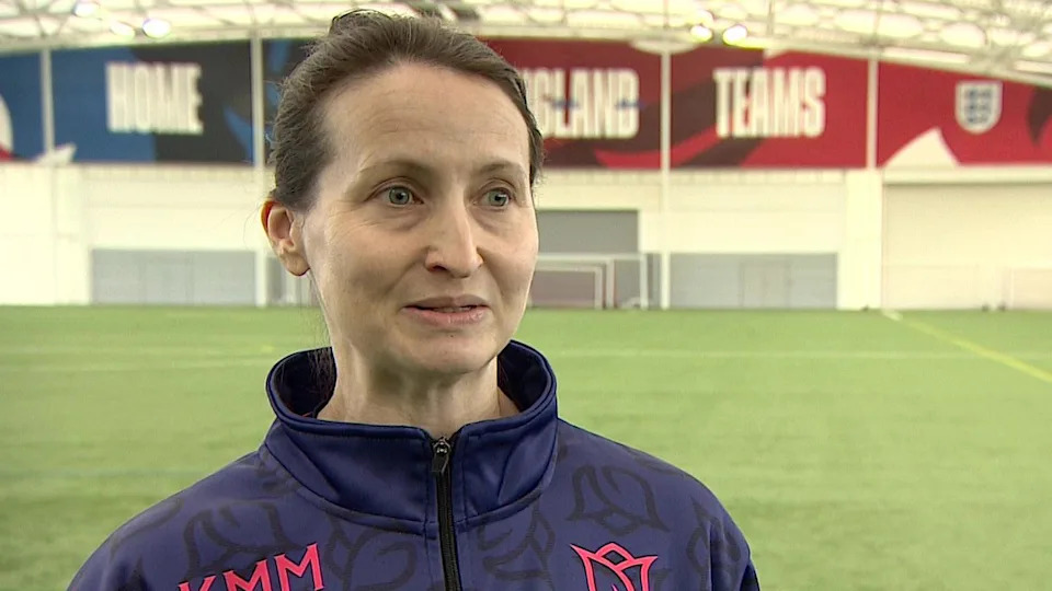 A women with brown hair tied up stood on a football pitch. She wears a blue top with some pink writing on it.