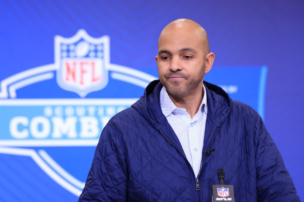 A man with a bald head and light stubble wearing a navy quilted jacket over a light blue patterned shirt speaks into an NFL Network microphone.