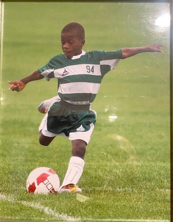 Jaffer Murphy as a child wears a green-and-white striped soccer uniform and kicks a red and white soccer ball on the field.
