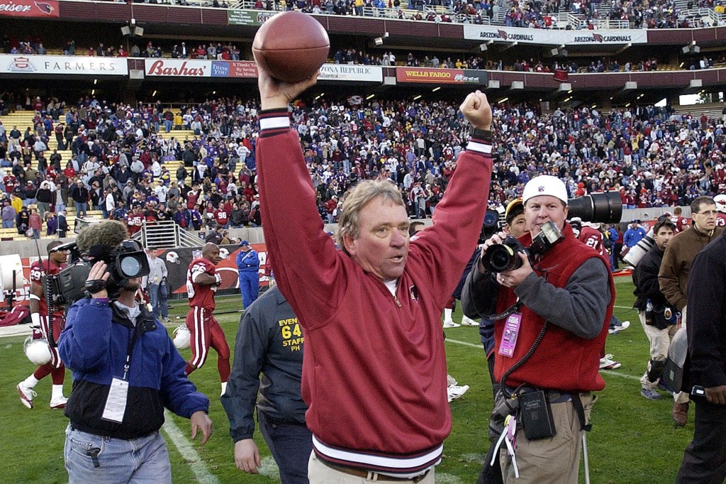 Arizona Cardinals head coach Dave McGinnis celebrates a win, raising a football in one hand and pumping his other fist in the air, on a football field filled with players and media.
