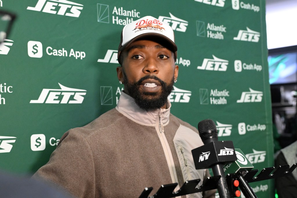 A man with a beard and a hat standing in front of a green banner with "JETS" and sponsor logos, speaking into a microphone.