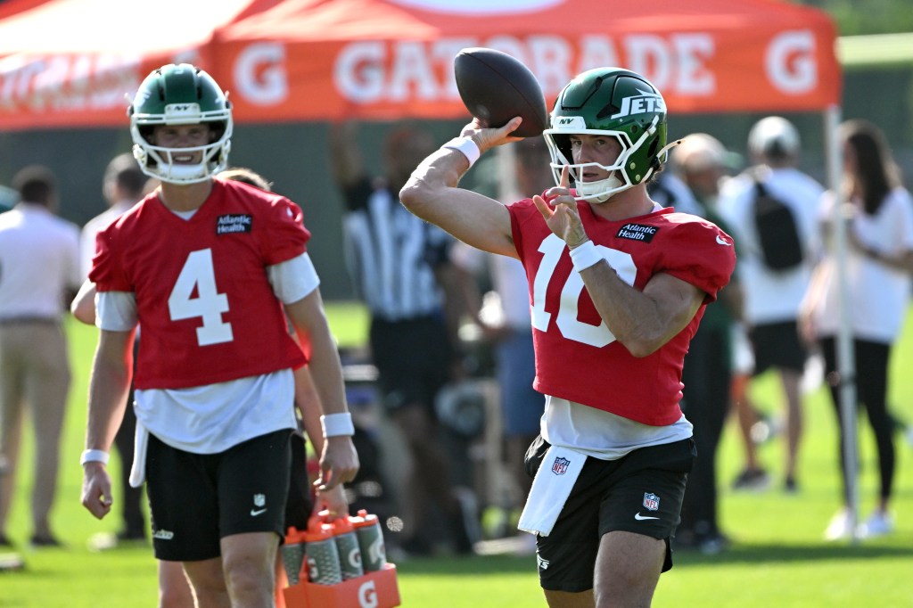 Zach Wilson in red practice jersey, green helmet and white wristbands, holds an NFL football in his right hand.