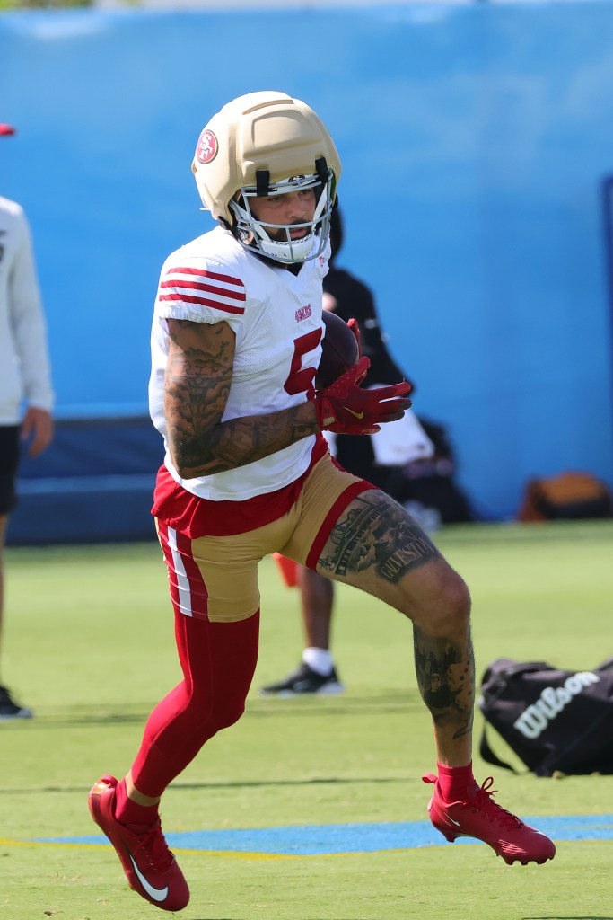 A San Francisco 49ers player in a white jersey and red pants, wearing a padded helmet, runs with a football during a joint practice.
