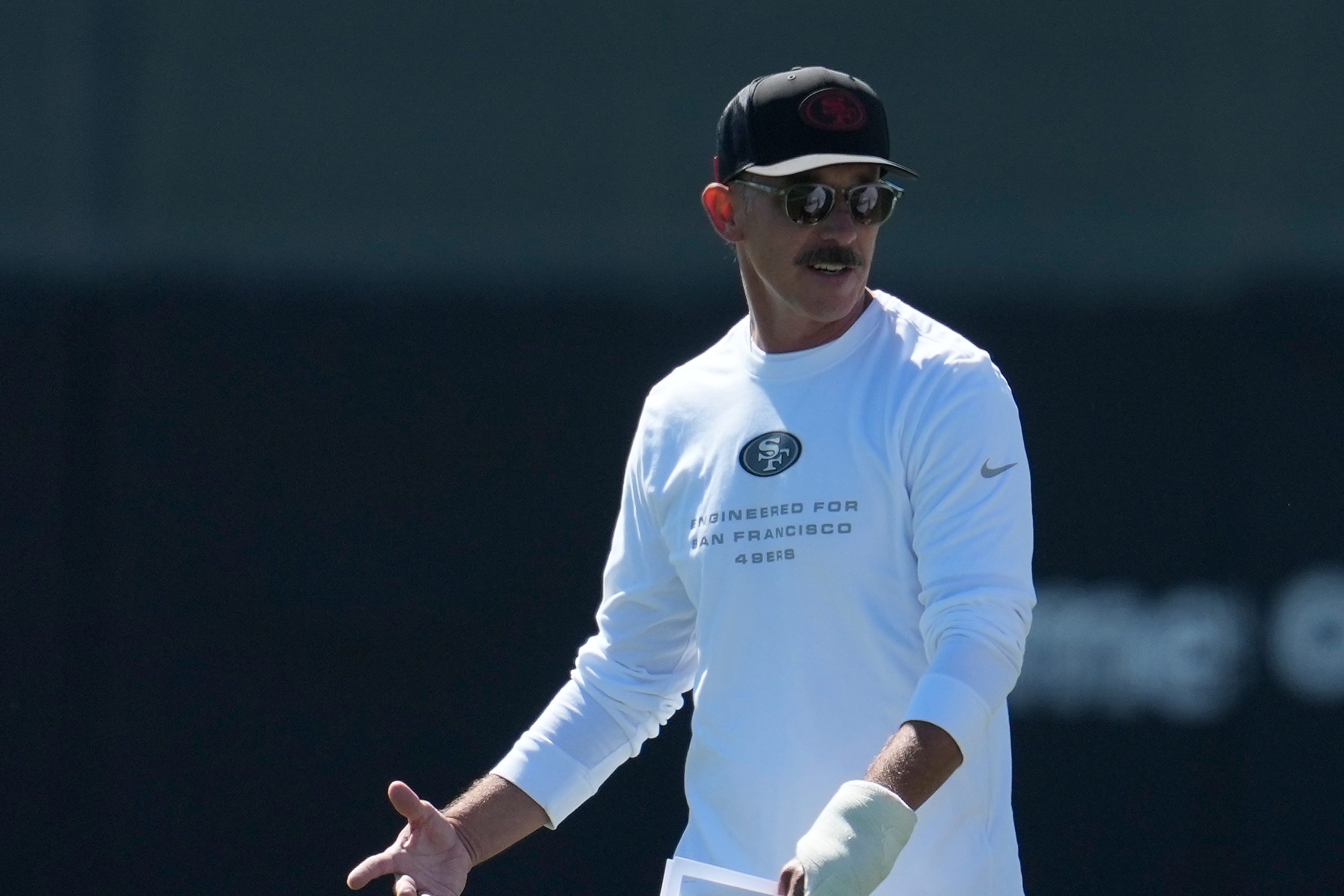 Kyle Shanahan, wearing a white long-sleeved shirt and black hat and sunglasses, talks to another coach.