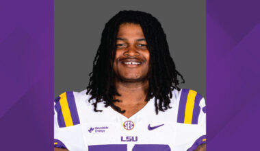 Portrait of a smiling LSU football player in a white jersey with purple and gold stripes and LSU logo, against a neutral backdrop.