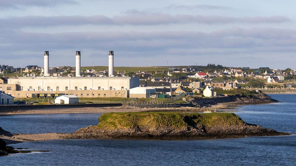 The power station is on the coast. It is a long box-shaped building with three chimneys.