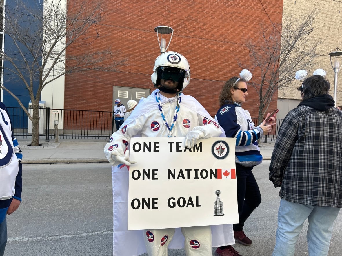 A man on a white suit and cape holds a sing and looks at the camera.