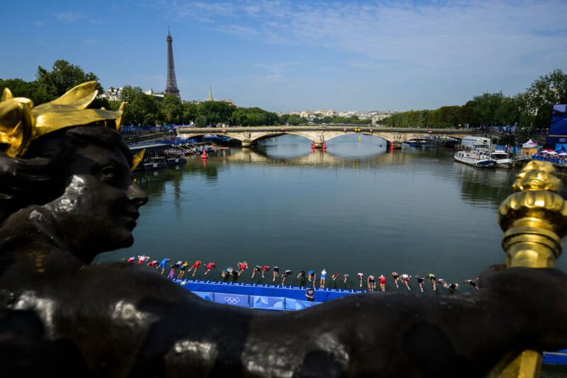 A bronze statue overlooks athletes diving into the Seine River during a swimming event in Paris, with the Eiffel Tower and a stone bridge visible in the background under a blue sky.