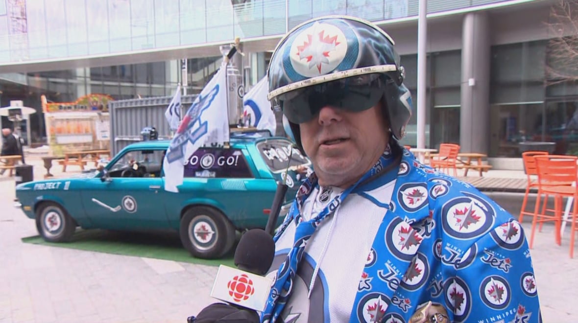 A man in a colourful blazer and fighter pilot helmet stands near a car decorated in Winnipeg Jets gear.
