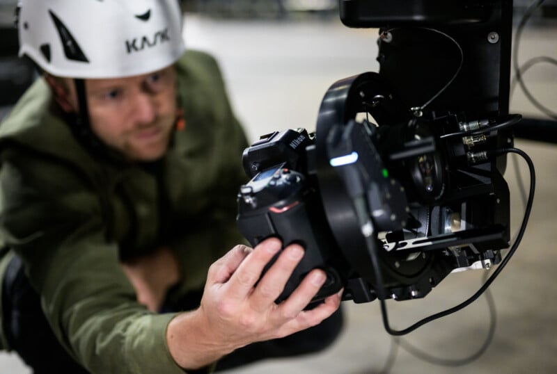 A person wearing a helmet and green jacket adjusts a professional camera mounted on specialized equipment, focusing intently on the camera's settings.