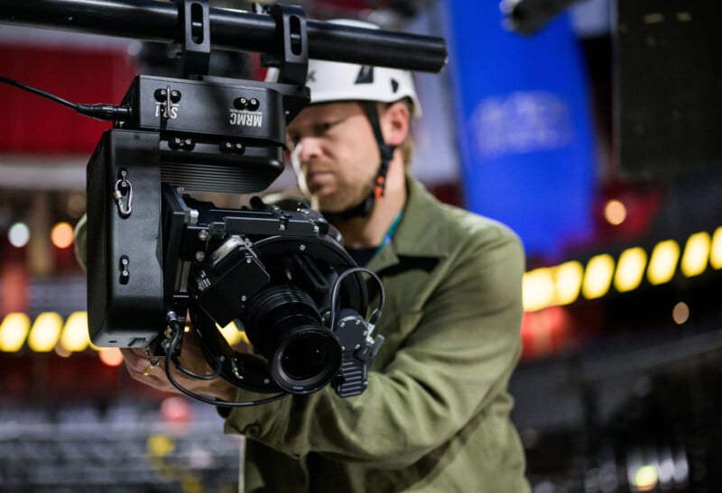 A person wearing a helmet adjusts a large professional video camera mounted on a rig, with blurred lights and a blue banner in the background.