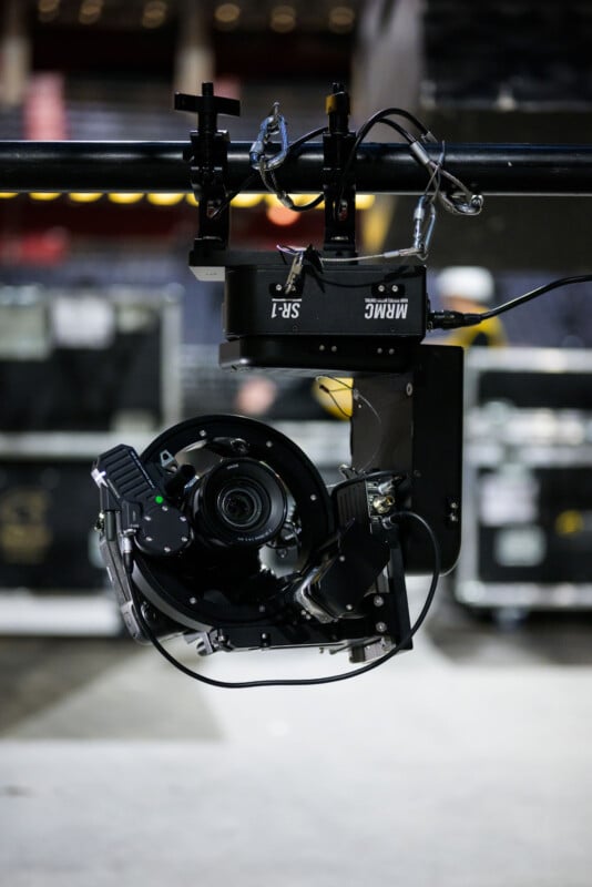 A close-up of a robotic camera rig labeled "MRMC SR-1" mounted on a rail system, with cables attached, set in an indoor venue with blurred equipment cases in the background.