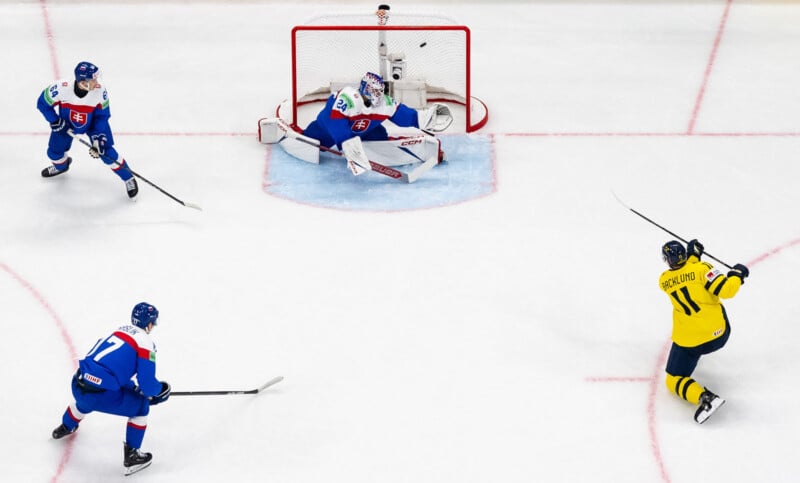 A hockey player in a yellow jersey prepares to shoot the puck at the goal, defended by a goalie and two players in blue jerseys on an ice rink.
