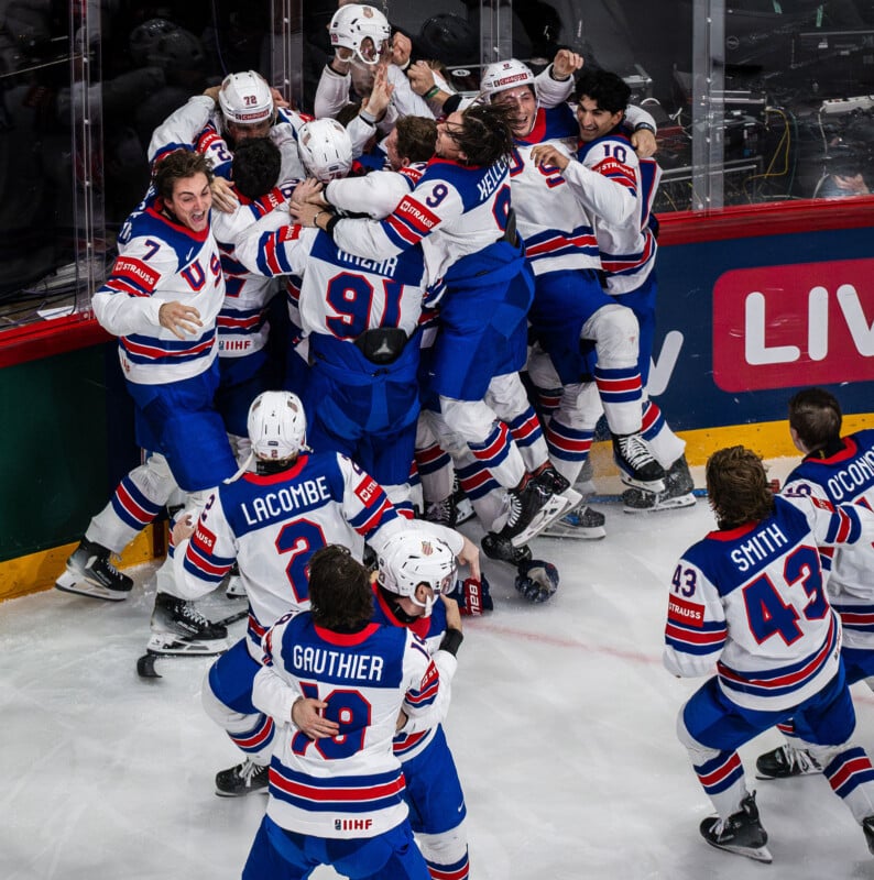 A group of ice hockey players in white, blue, and red uniforms joyfully huddle and celebrate near the rink boards, with some players jumping and others raising their arms in excitement.