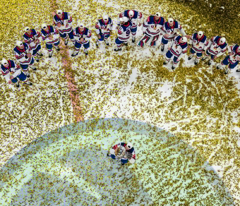 A hockey team in white, blue, and red uniforms stands in a semicircle on an ice rink covered in gold confetti, watching two teammates holding a trophy at the center.