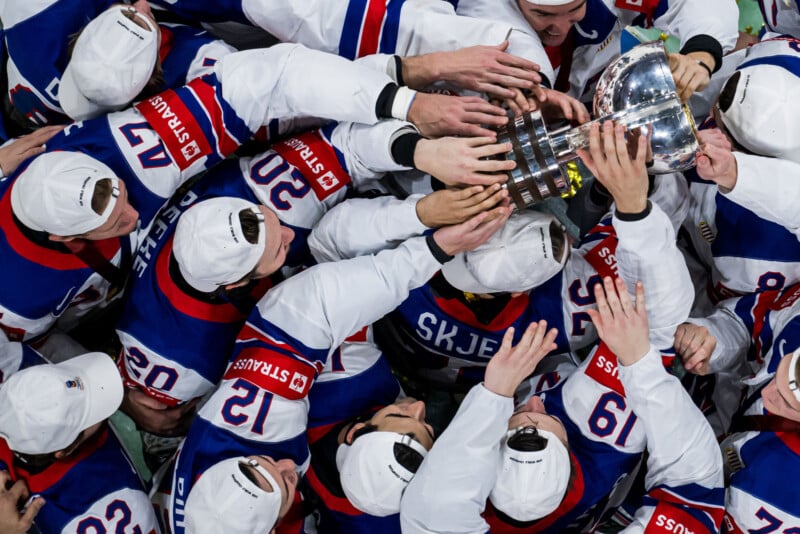 A group of ice hockey players in blue, red, and white uniforms and white caps joyfully reach out together to touch and lift a silver trophy, celebrating their victory.