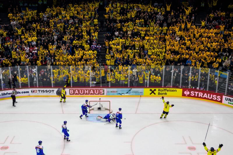 A hockey player in yellow celebrates a goal as the opposing blue team stands near the net; behind the glass, a large crowd of fans in yellow jerseys cheer in a packed arena.
