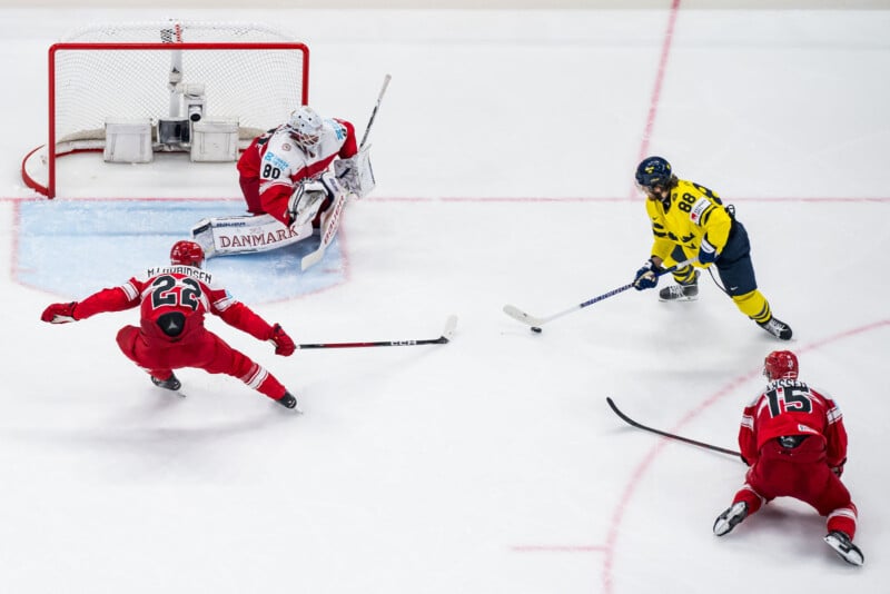 A Swedish ice hockey player in yellow approaches the Danish goal, aiming to shoot, while the Danish goalie and two defenders in red try to block his attempt on the ice rink.