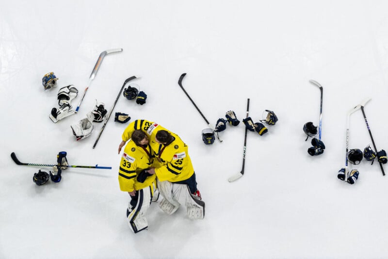 Two hockey players in yellow jerseys embrace on the ice, surrounded by scattered sticks, gloves, and helmets, suggesting a celebration after a game. The white ice offers a stark background to the scene.